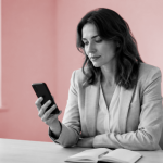 Woman in a gray blazer in a bright white office studies her phone, illustrating real-time Wangiri fraud detection in action.